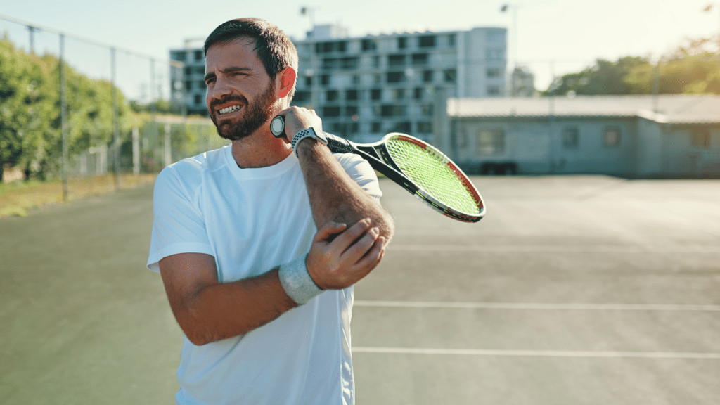Man holds his elbow in pain holding a tennis racket, experiencing tennis elbow and in need of help from a physical therapist