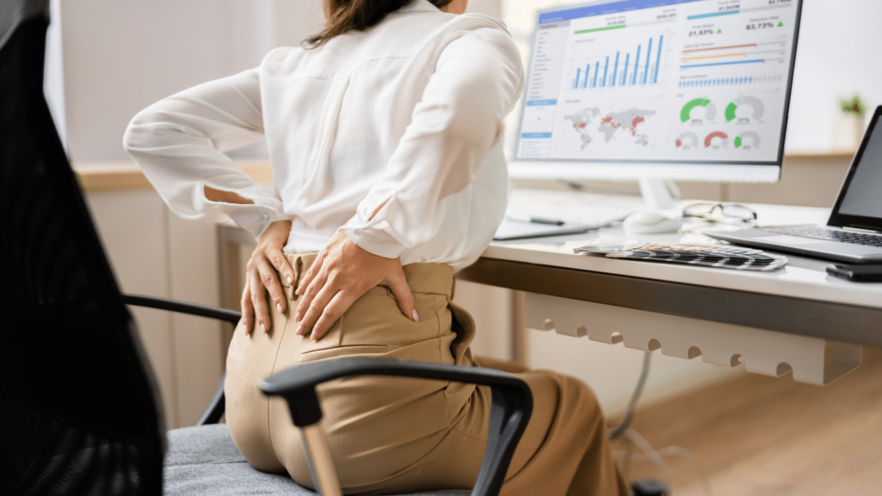 Woman holds back in pain while sitting at desk, wondering How Your Desk Job Is Weakening Your Hips