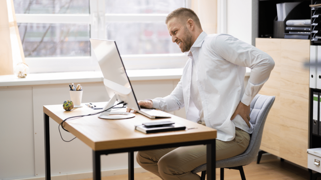 Man holds back in pain while sitting at desk, wondering How Your Desk Job Is Weakening Your Hips